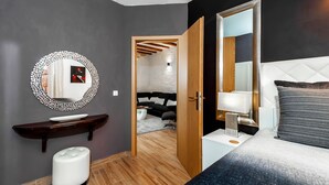 Ein modernes Schlafzimmer mit einem großen Bett, einem Nachttisch mit Lampe, einem runden Spiegel mit dekorativem Rahmen, einem kleinen weißen Hocker und einer Holzbank.
