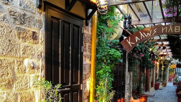 A narrow cobblestone street with a stone building featuring a black door and a yellow awning. There are potted plants and a hanging bird feeder. - Grand Master Suite (Rodos)