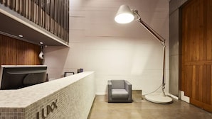Un área de recepción con un mostrador identificado con 'LUX', una silla gris, una lámpara de pie y una puerta de madera.