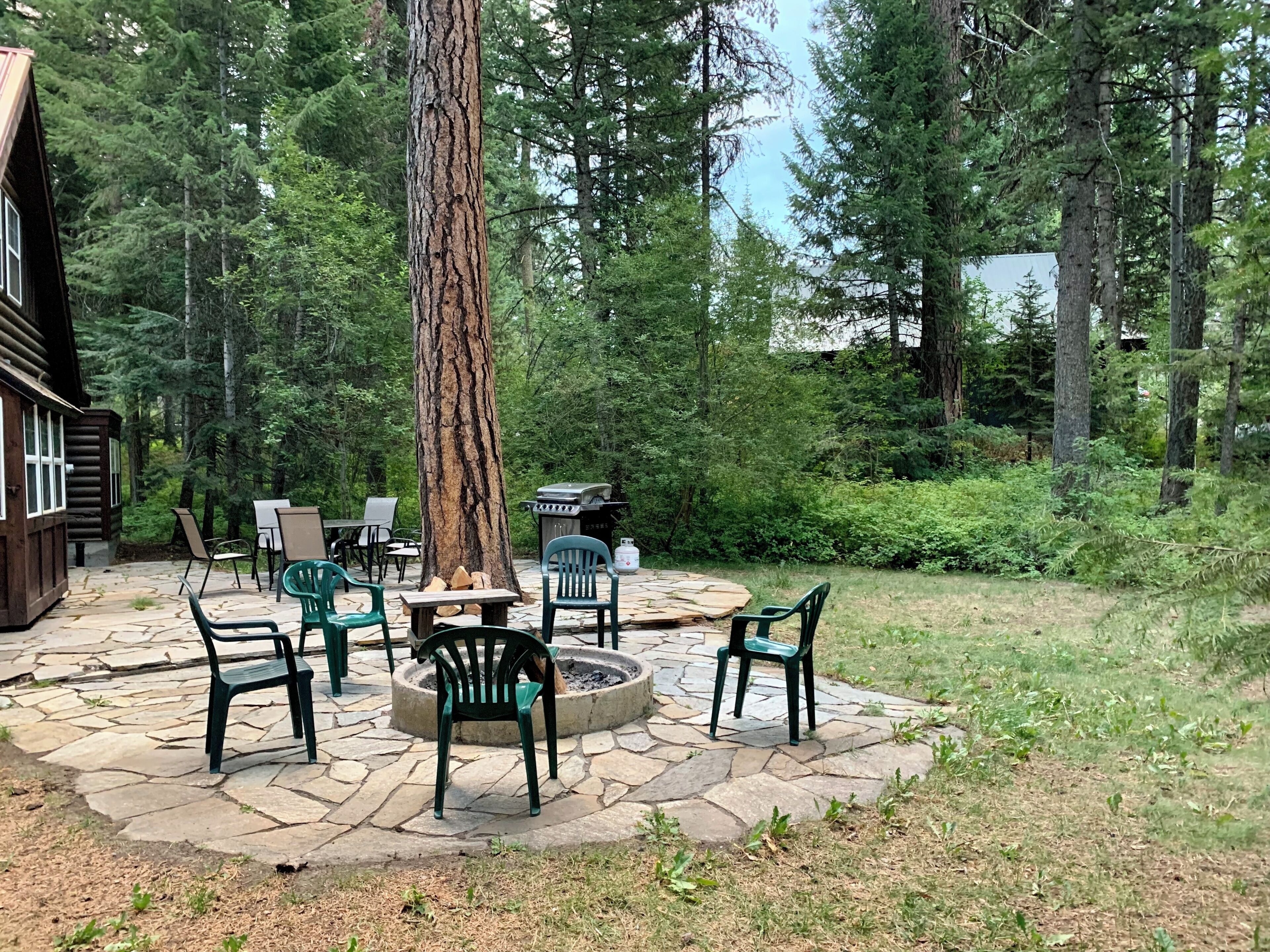 A stone patio with outdoor seating and a fire pit, surrounded by a forested area.
