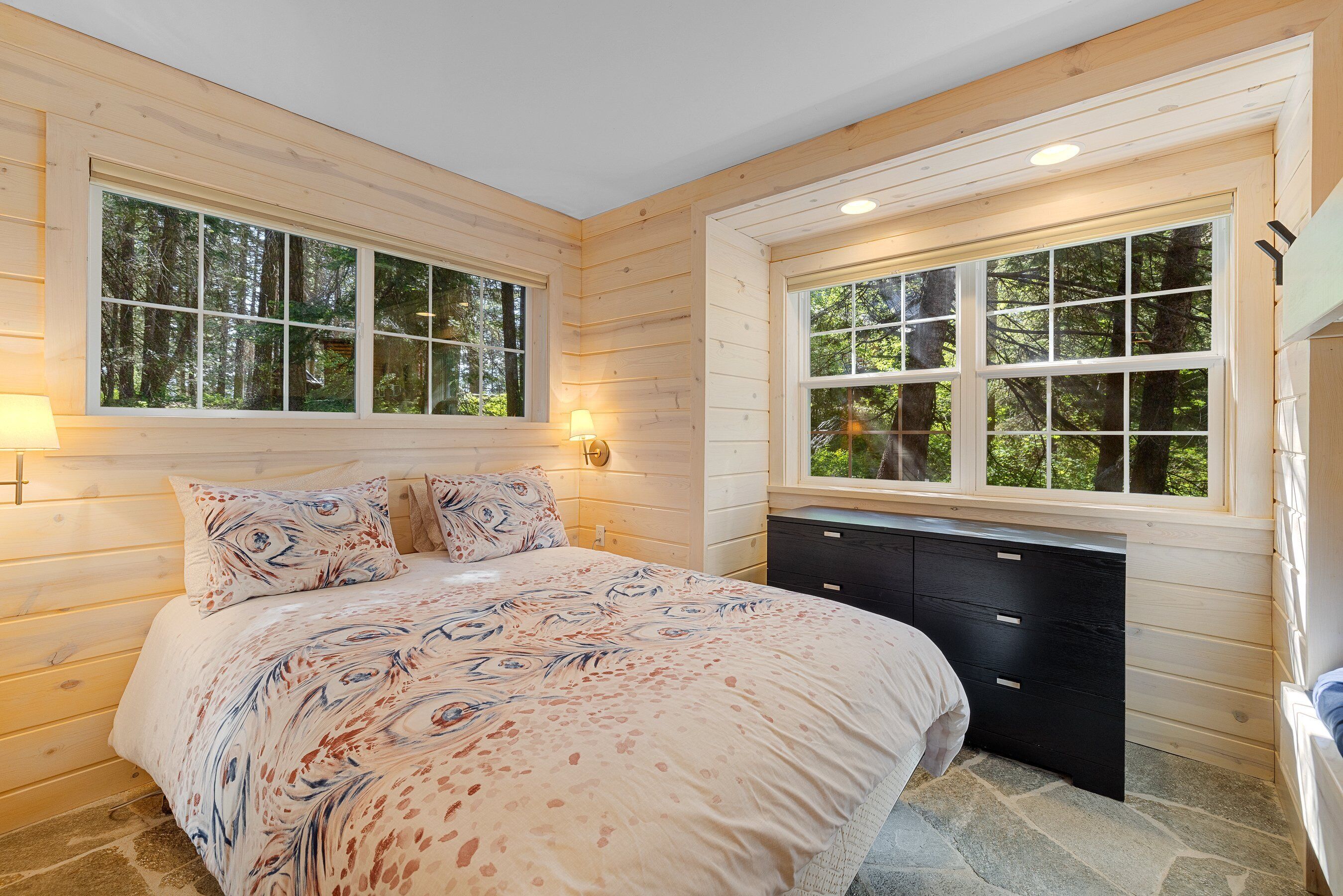 A bedroom with a large bed, two bedside lamps, a dresser, and a window with a view of trees.
