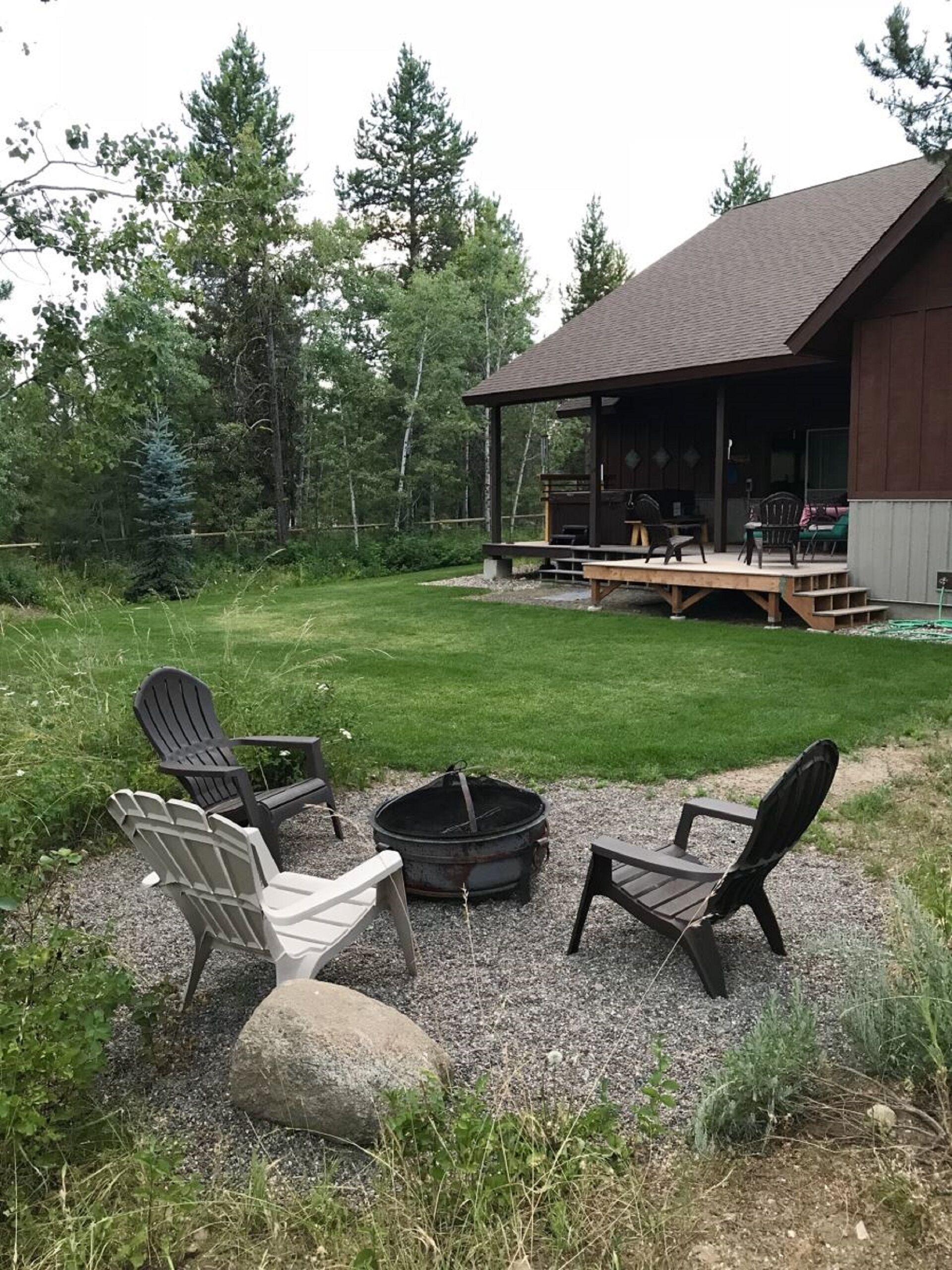 A covered outdoor seating area with Adirondack chairs and a fire pit, surrounded by a grassy lawn and trees.