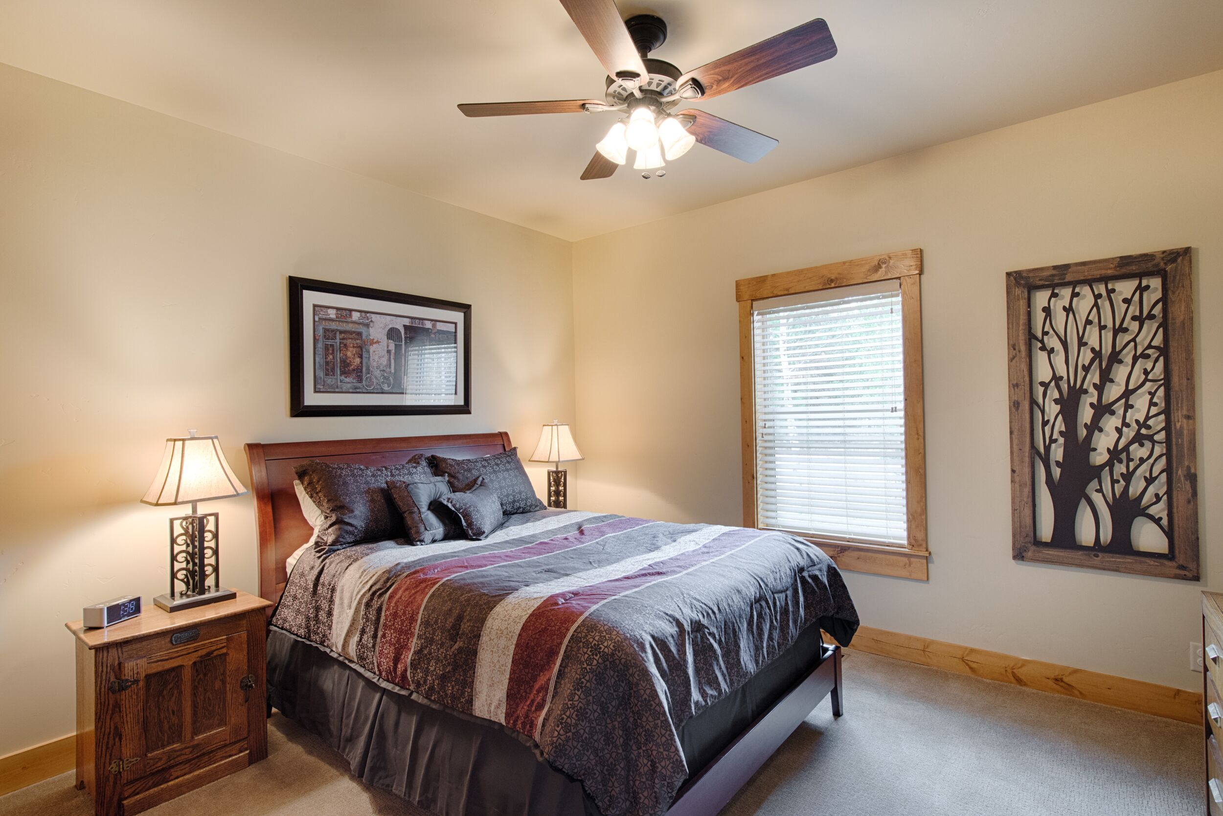 A bedroom with a bed, two bedside tables with lamps, a ceiling fan, a window with blinds, and a wall decoration.