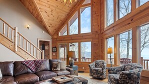 Un interior de cabaña espacioso con escalera de madera, muebles de cuero y grandes ventanales que ofrecen vistas al exterior.