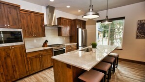 A modern kitchen with wooden cabinets, stainless steel appliances, a large island with bar stools, and a window providing natural light. - Suite Retreat: Short Walk to Downtown McCall, A/C, Propane Fireplace (McCall)