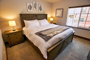 Quarto com cama de madeira, roupa de cama branca, manta decorativa, abajures e arte na parede.