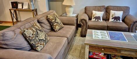 A living room with two brown leather sofas, patterned cushions, a wooden coffee table with books, and a lamp.