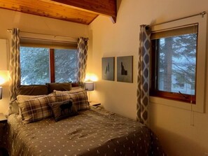 A cozy bedroom with a bed, two bedside lamps, and a window with curtains. There are two framed pictures on the wall.
