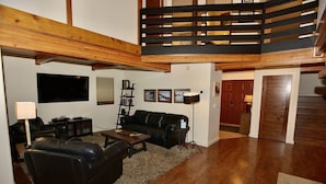 Ein geräumiges Wohnzimmer mit einer schwarzen Ledersofa, einem Holztisch und einem großen Flachbildfernseher an der Wand. Eine Holztreppe mit schwarzer Geländer führt auf eine obere Ebene.