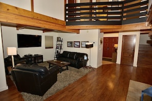A spacious living room with a black leather sofa, a wooden coffee table, and a large flat-screen TV mounted on the wall. A wooden staircase with a black railing leads to an upper level. - Fairway Lodge: Beautifully Updated, On the Golf Course, Gas Fireplace (McCall)