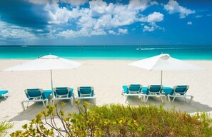 A beach with white sand, turquoise water, and clear skies. There are rows of blue lounge chairs under white umbrellas.