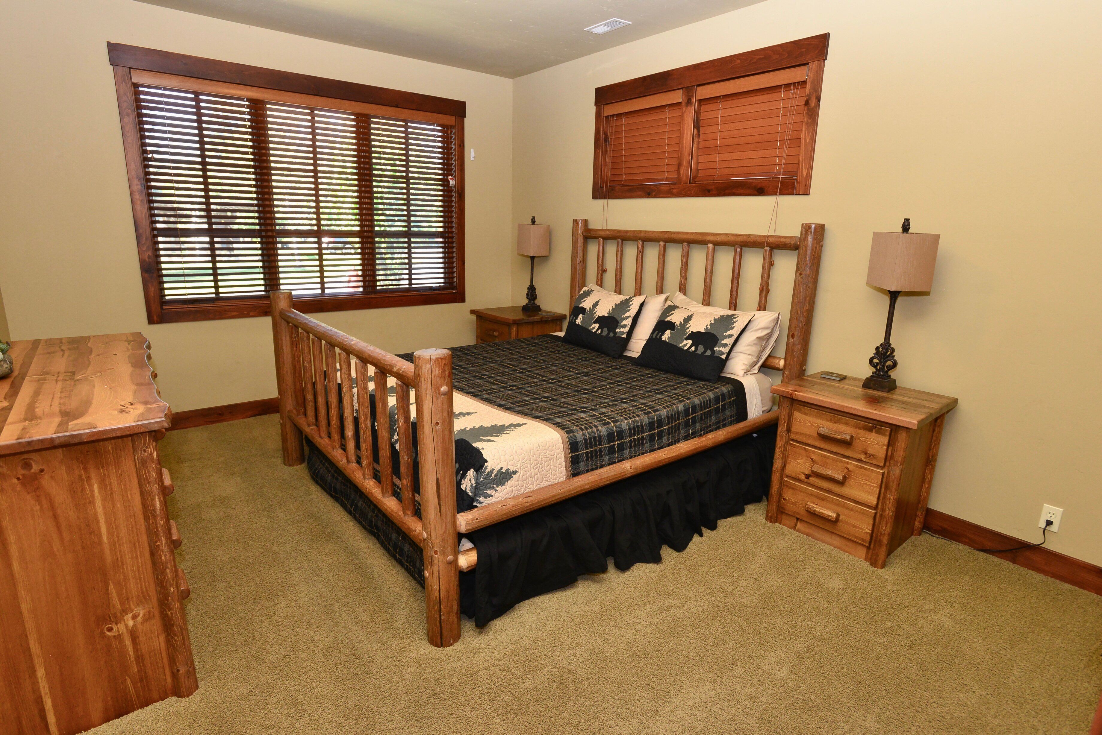 A bedroom with a wooden bed, two bedside tables with lamps, a wooden dresser, and a window with blinds.