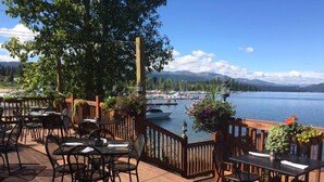 An outdoor dining area with tables and chairs, overlooking a lake with boats and mountains in the background.
