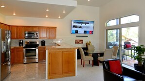 A modern kitchen with wooden cabinets, stainless steel appliances, and a large island. There is a dining area with a table and chairs, and a TV mounted on the wall.