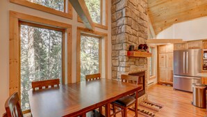 Un comedor con mesa y sillas de madera, ventanas amplias con vistas al bosque, chimenea de piedra y cocina equipada con electrodomésticos modernos.