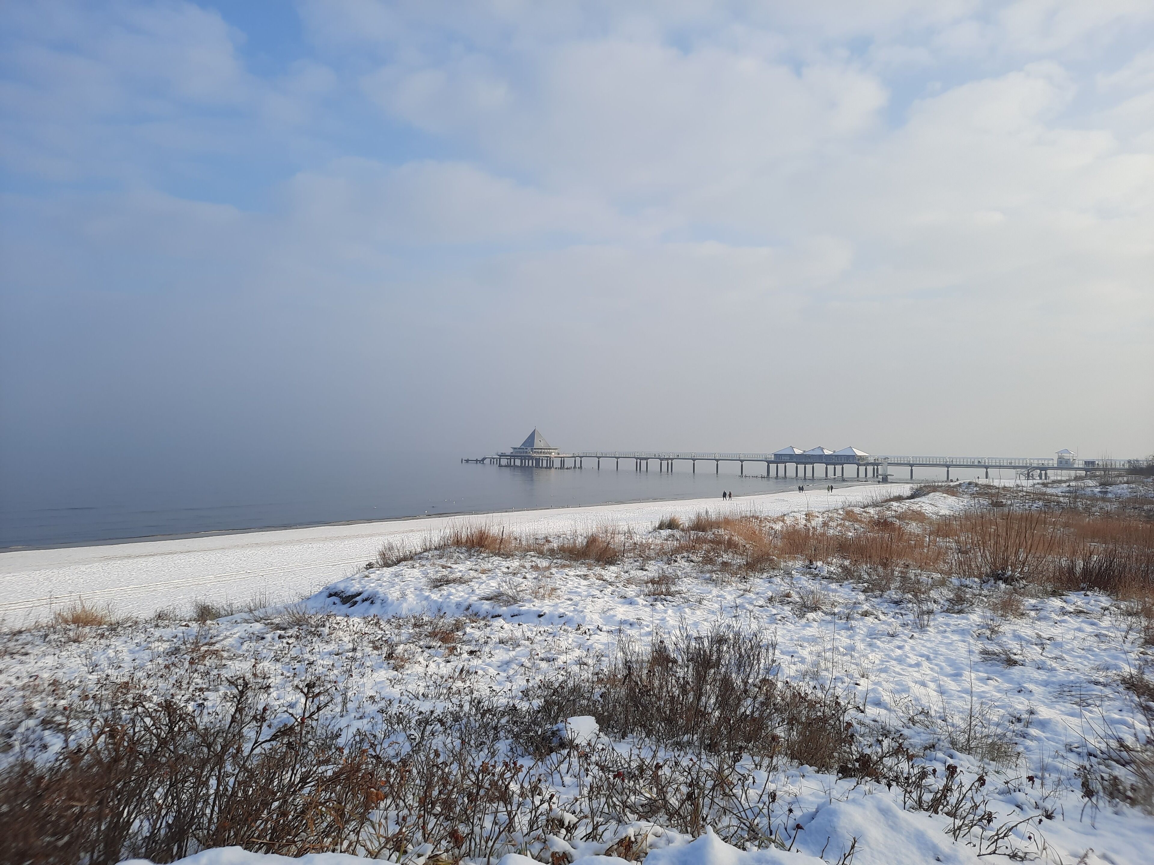 Een kustlandschap met een pier die in het water steekt, een zandstrand en een met sneeuw bedekte grond.