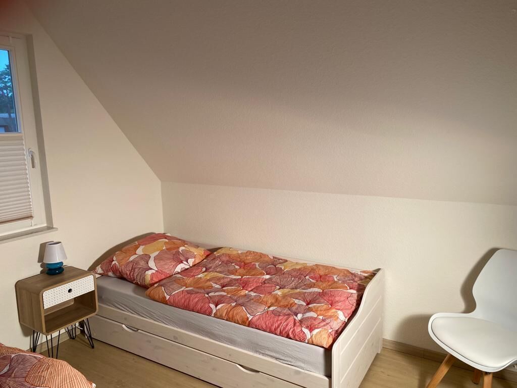 Een kleine kamer met een enkel bed, een houten hoofdbord, een nachtkastje met lamp en een witte stoel.