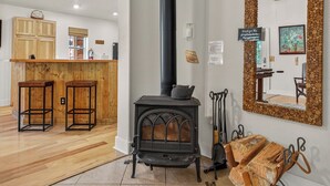 A room with a wood-burning stove, a kitchen area with wooden cabinets, and a large mirror with a stone frame. - Hubbard Cabin in McCall - near Payette Lake, private hot tub, quiet (McCall)