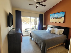 A hotel room with a bed, a TV, a ceiling fan, a painting, and a view of the outdoors. - Condo in Pedregal/3 King beds/2 pools/Walk to town/Views (Cabo San Lucas)