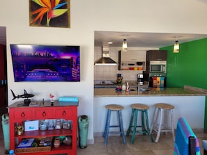 A modern kitchen with a green accent wall, a flat-screen TV, and a bar area with stools. - Condo in Pedregal/3 King beds/2 pools/Walk to town/Views (Cabo San Lucas)