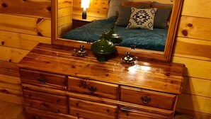 Un comò di legno con uno specchio sopra, che riflette un letto con cuscini e una lampada.