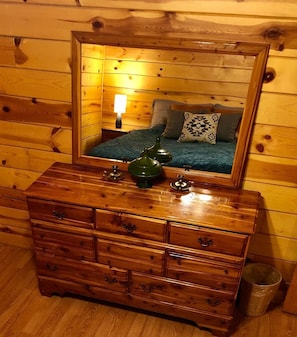 Un aparador de madera con un espejo arriba, que refleja una cama con almohadas y una lámpara.