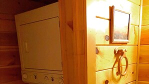 Una stanza compatta in stile baita, con arredamento in legno, dotata di lavatrice, un piccolo lavandino e un armadietto in legno con portasciugamani.