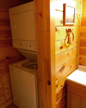Una habitación tipo cabaña, de tamaño compacto, con lavadora, un lavamanos pequeño y un mueble de madera con un perchero para toallas.