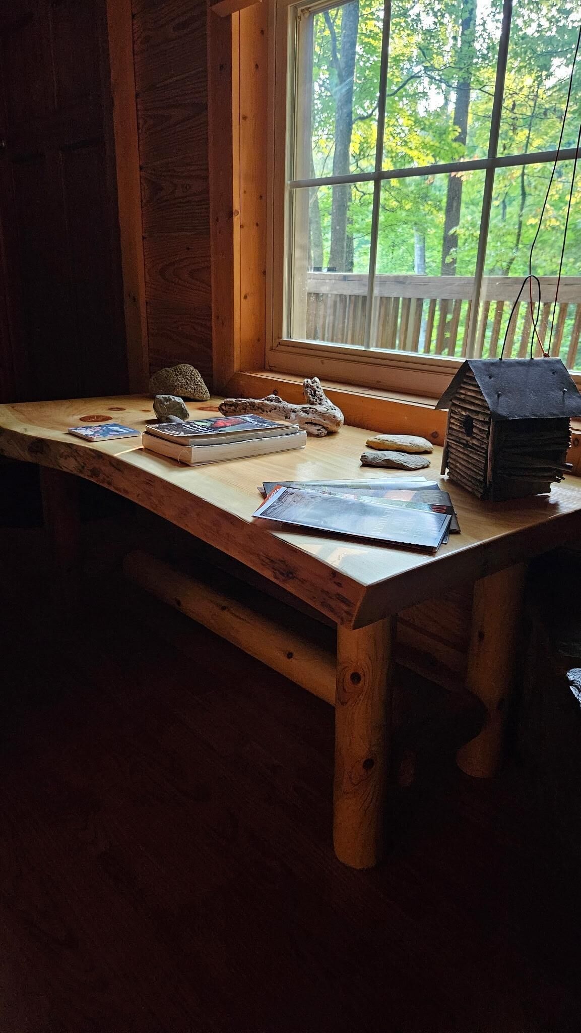 A wooden table with books and a birdhouse in a cabin with a window overlooking a forest.