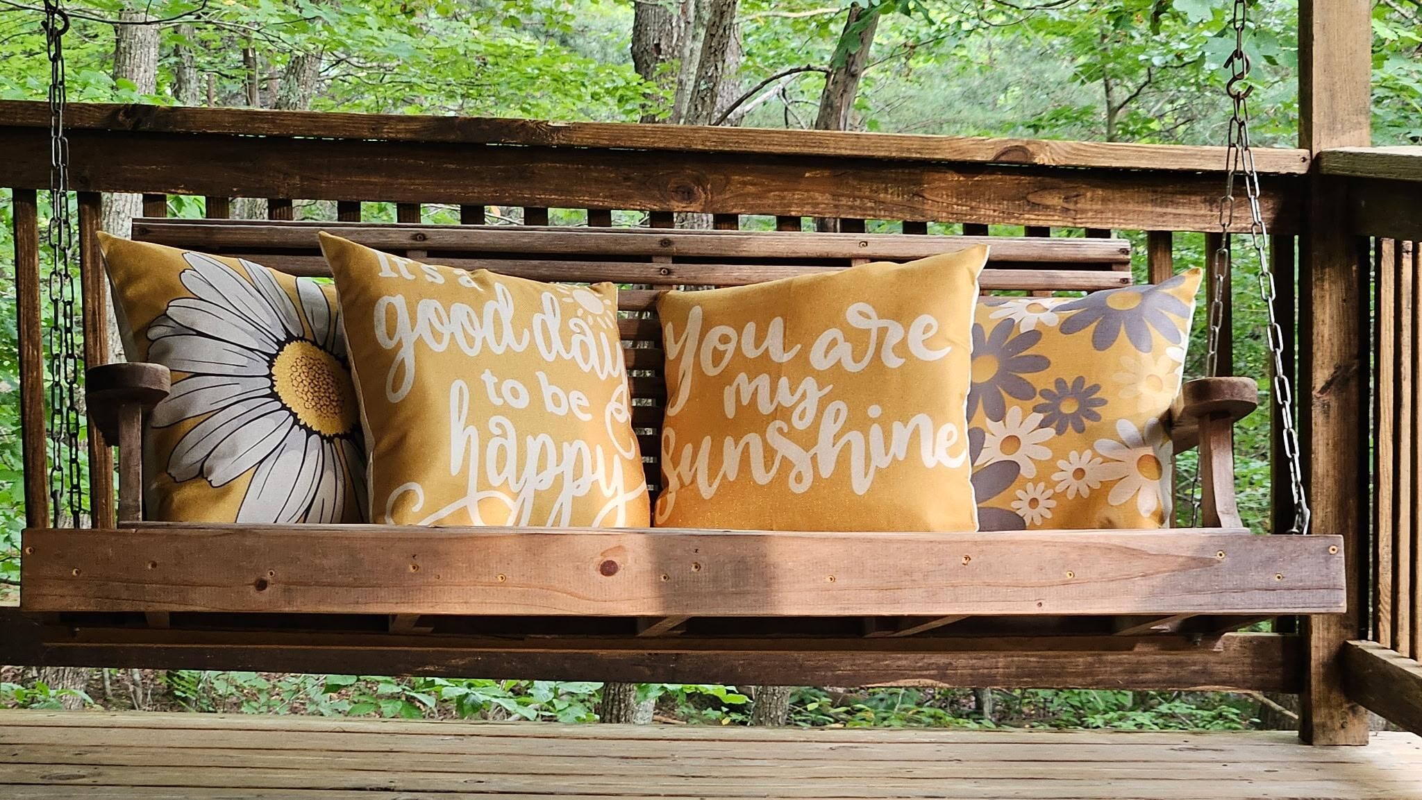 A wooden swing bench with three cushions, one with a daisy design and two with motivational text.
