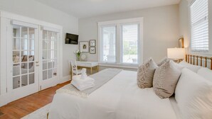 A bedroom with a large bed, a desk, and a chair. There are white doors and windows with blinds. - The Pink Door - 8th Ave Beauty - Stunning & Fresh (Nashville)
