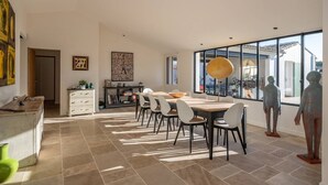 A spacious dining area with a large table and chairs, a kitchen area with cabinets and a countertop, and a large window offering a view of the outside. - Arte - Artist villa in the heart of la Noue (Sainte-Marie-de-Ré)