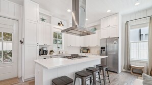 A modern kitchen with white cabinetry, a large island with stools, a stainless steel range hood, and a built-in refrigerator.