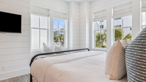 A bedroom with a bed, a TV mounted on the wall, and large windows offering a view of palm trees and buildings.