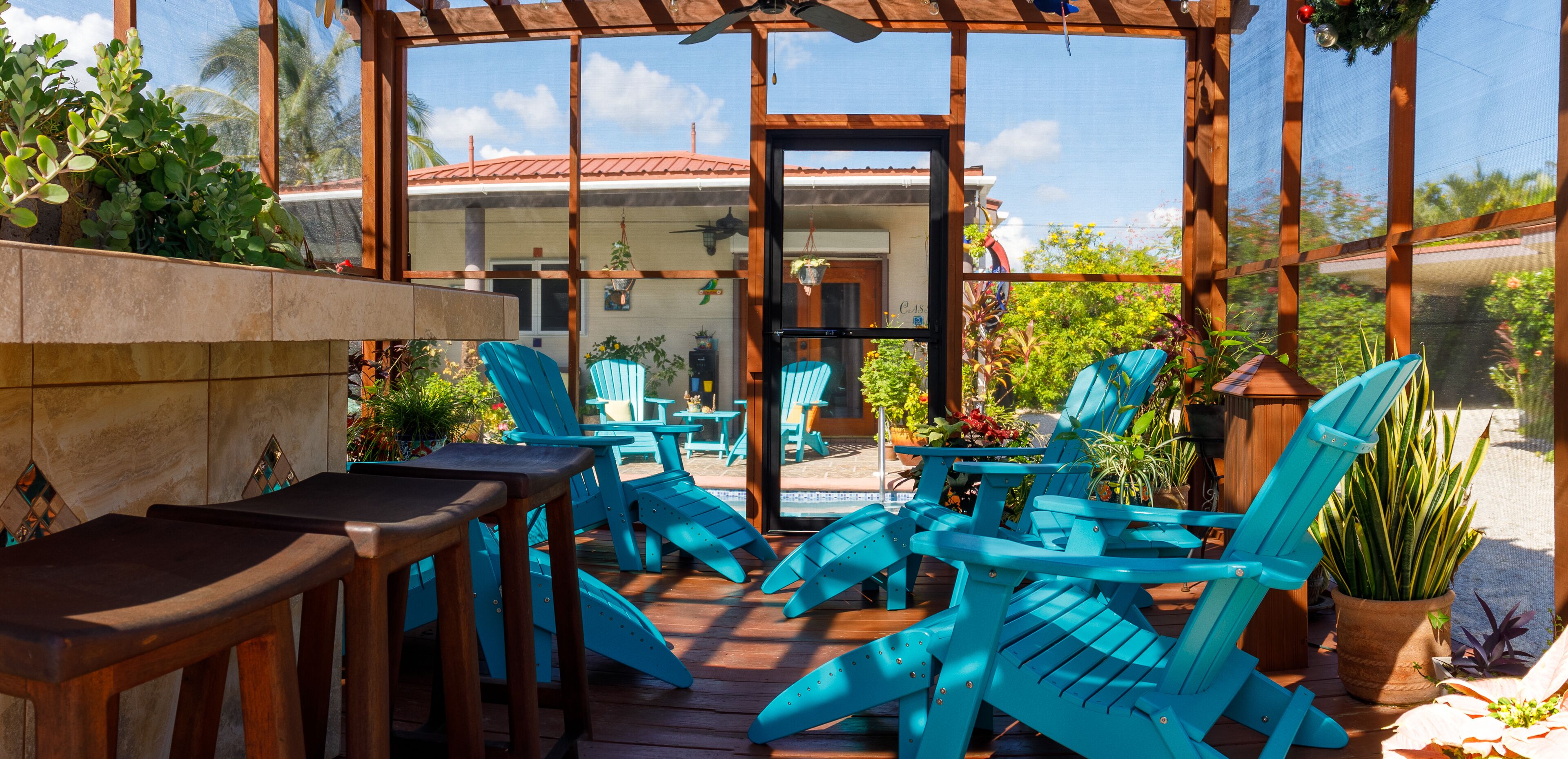A covered outdoor seating area with wooden tables and blue chairs, surrounded by potted plants and a view of a house with a red roof.