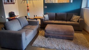 A living room with a grey sofa, a brown ottoman, and a patterned rug. - Mountain Side Forest Retreat - Hot Tub!! - 1br 1ba <3beds> (Anchorage)