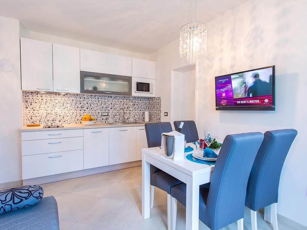 A modern kitchen with white cabinets, a microwave, and a tiled backsplash. There is a dining table with blue chairs and a TV mounted on the wall displaying a program.