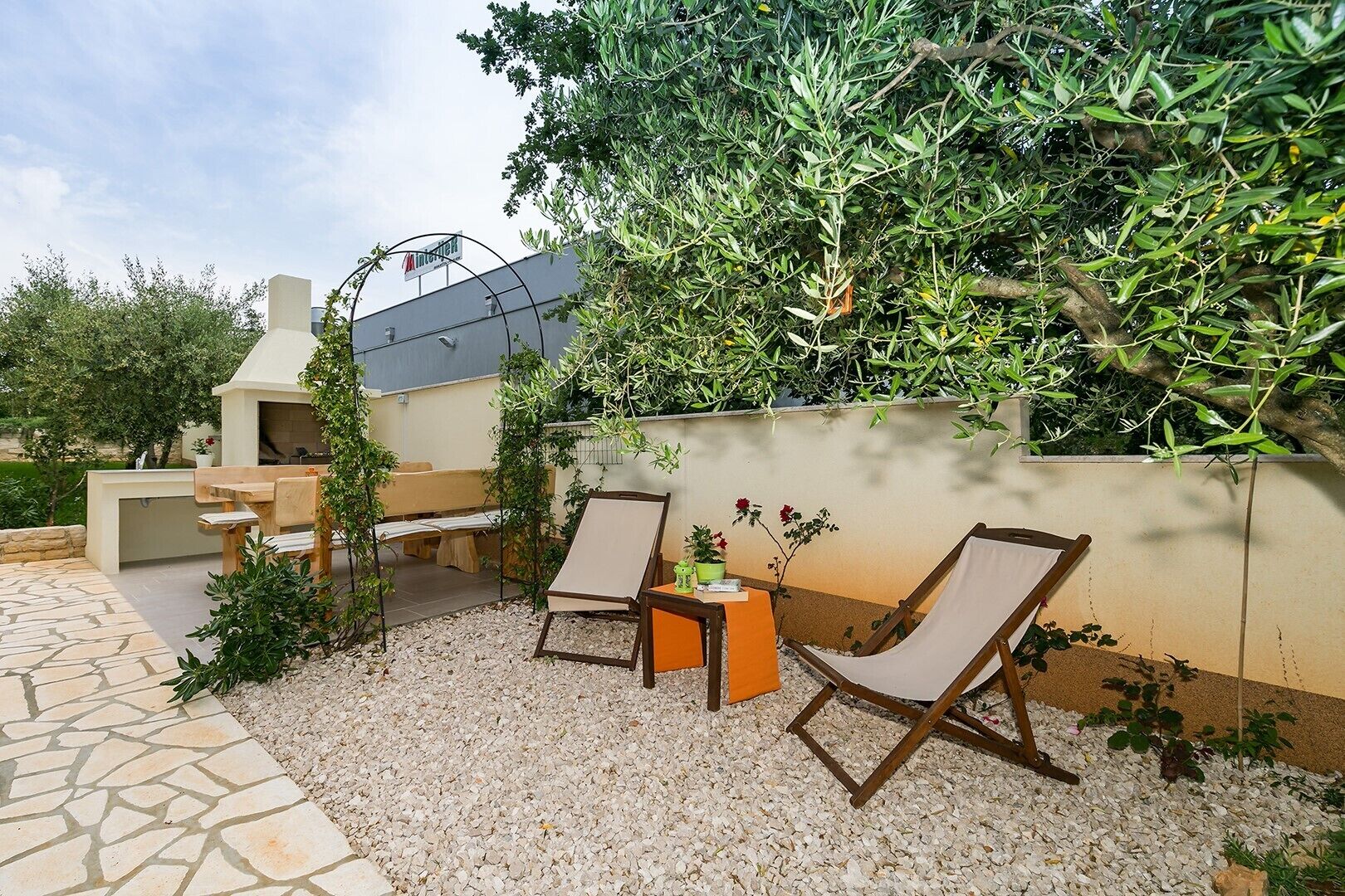 Een buitenpatio met een stenen vloer, twee houten lounge fauteuils, een bijzettafeltje en een barbecuehoek.