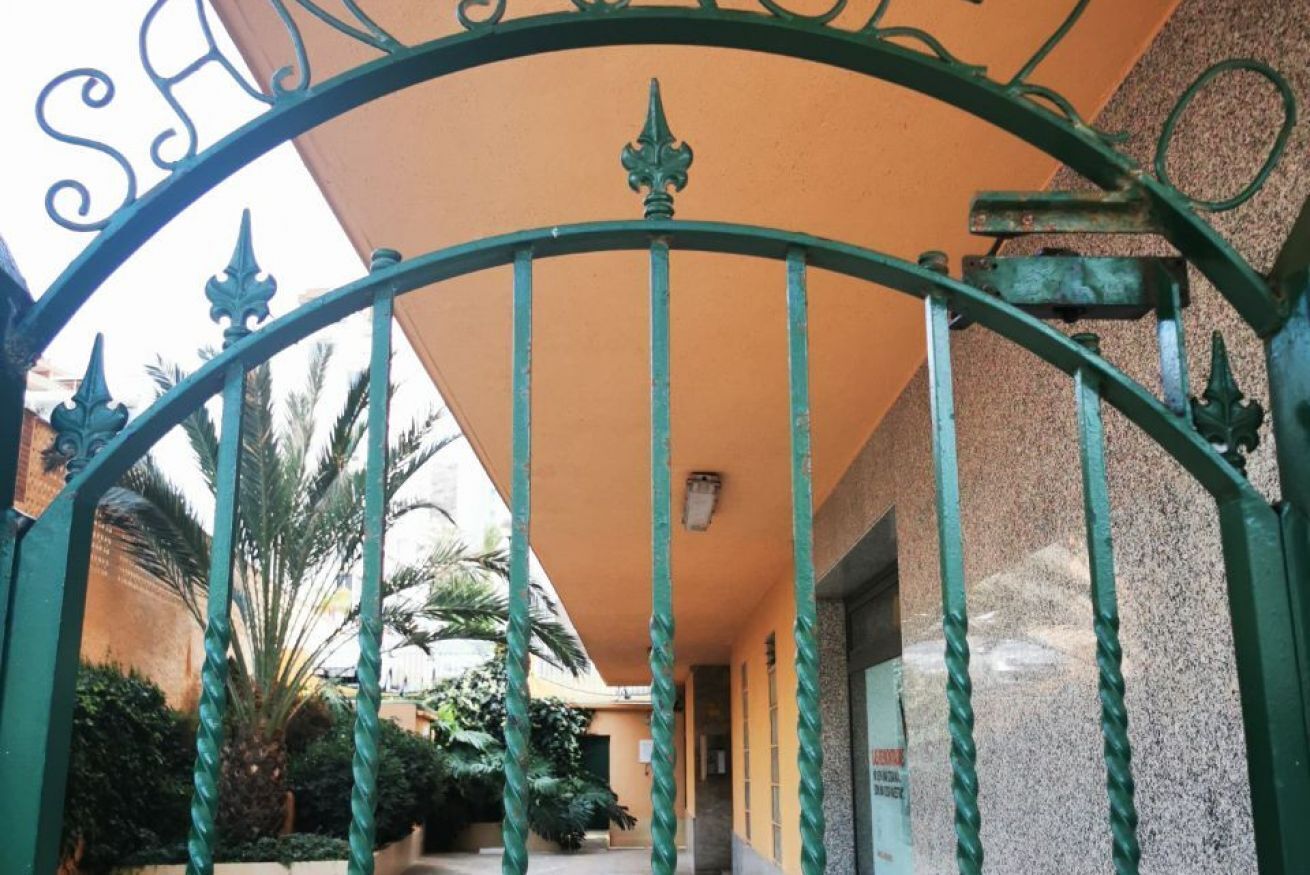 A wrought iron gate with a decorative top, leading to a building with a beige facade and a covered walkway.