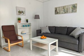 A living room with a grey sofa, a brown chair, a white coffee table with oranges, a floor lamp, and a wall with a Buddha painting.