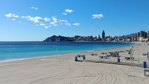 A beach with a city skyline in the background, clear skies, and a sandy shore with blue chairs and trash bins. - SAN ANGELO Levante beach apartments (Benidorm)