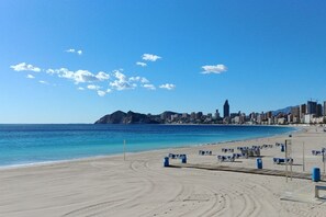 Una playa con un horizonte urbano al fondo, cielos despejados y una orilla arenosa con sillas azules y botes de basura.