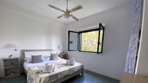A bedroom with a bed, bedside tables, a ceiling fan, a window with an open shutter, and a view of trees outside. - Apartment Ionian coast (Sant'Andrea Apostolo dello Ionio)