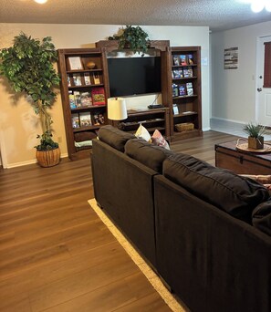 A living room with a sectional sofa, a flat-screen TV, wooden bookshelves, and a potted plant. - Peaceful, Cozy & Spacious Lower Level Dwelling (Bella Vista)