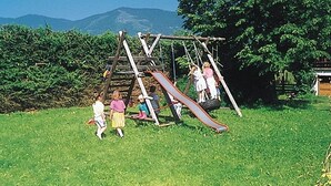 A playground with a wooden swing set and slide in a grassy area with trees and mountains in the background. - Pferdegut Romerhof (Altenmarkt im Pongau)