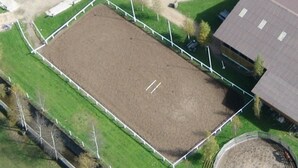 An aerial view of a fenced equestrian arena with a dirt surface, surrounded by green grass and a building with a sloped roof. - Pferdegut Romerhof (Altenmarkt im Pongau)