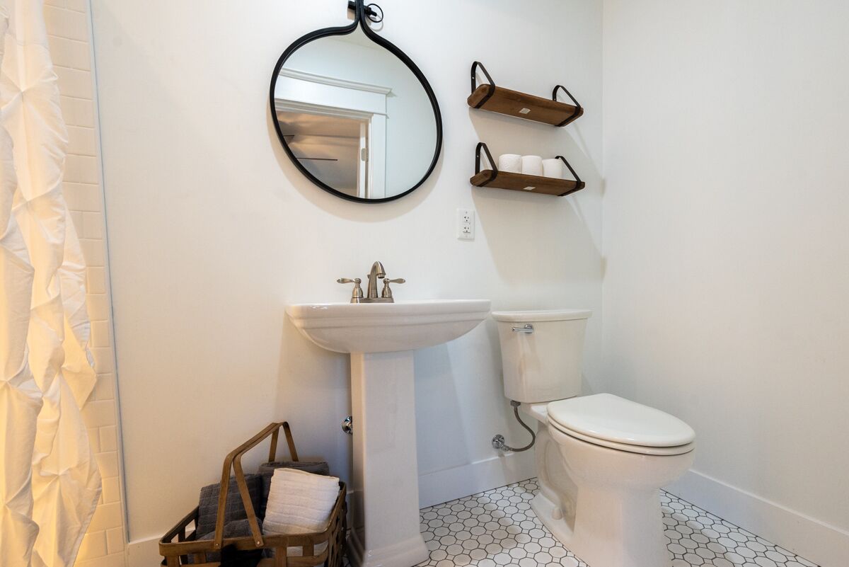 Et badeværelse med en hvid vask, toilet og et rundt spejl. Der er træhylder over toilettet og en kurv med håndklæder på gulvet.