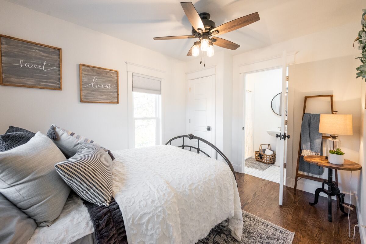 A bedroom with a bed, a ceiling fan, a bedside table with a lamp, and a door leading to another room.