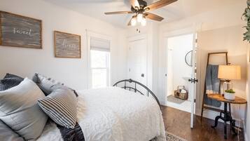 A bedroom with a bed, a ceiling fan, a bedside table with a lamp, and a door leading to another room.