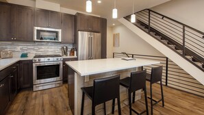 A modern kitchen with dark wood cabinets, stainless steel appliances, a central island with bar stools, and a staircase with a metal railing. - Arrow at Winter Park Unit 248 Downtown 4 bedroom private hot tub & mountain view (Winter Park)